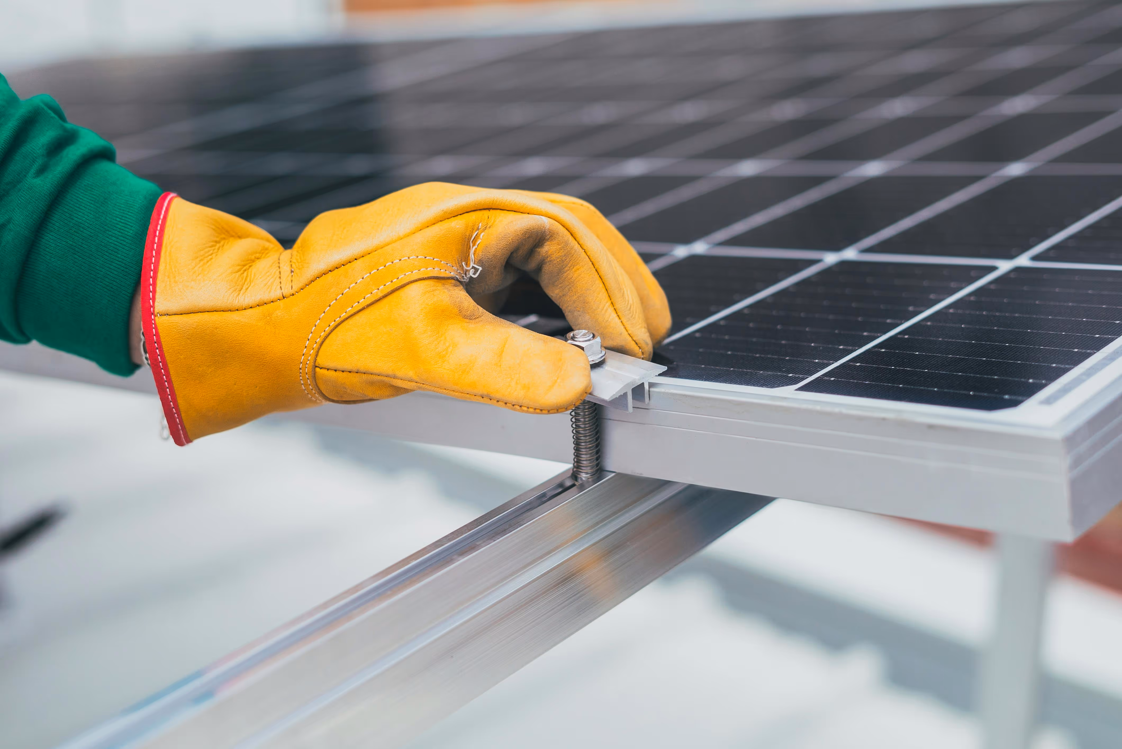 Hand wearing a yellow work glove adjusting a bolt on a solar panel mounting bracket.