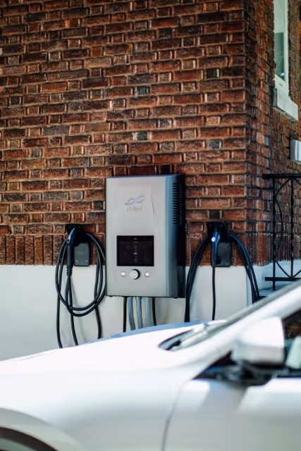 Electric vehicle charging station mounted on a brick wall with charging cables and a white car partially visible in the foreground.