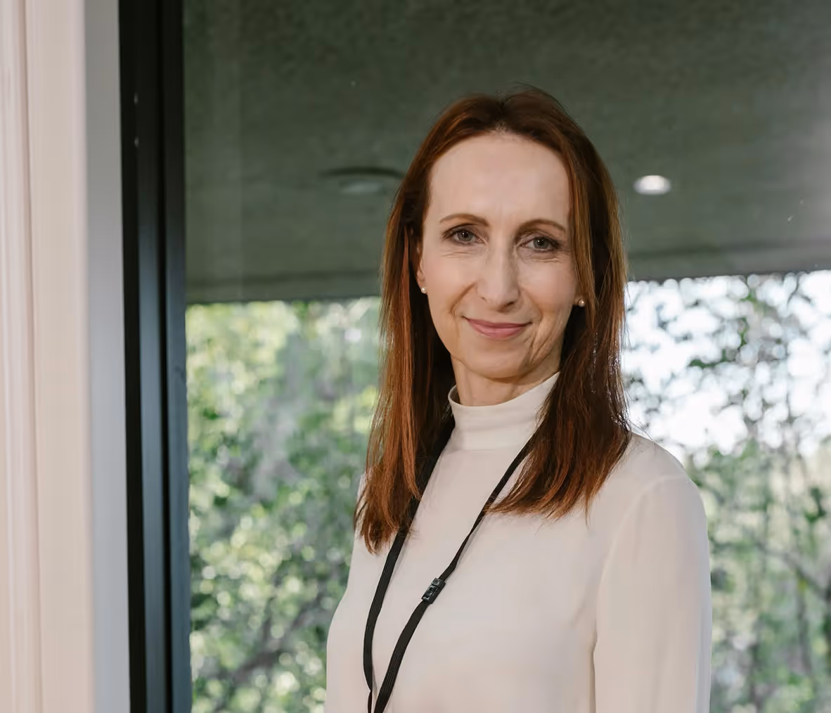 Woman with straight brown hair wearing a white turtleneck and black lanyard, smiling gently in front of a window with green trees outside.