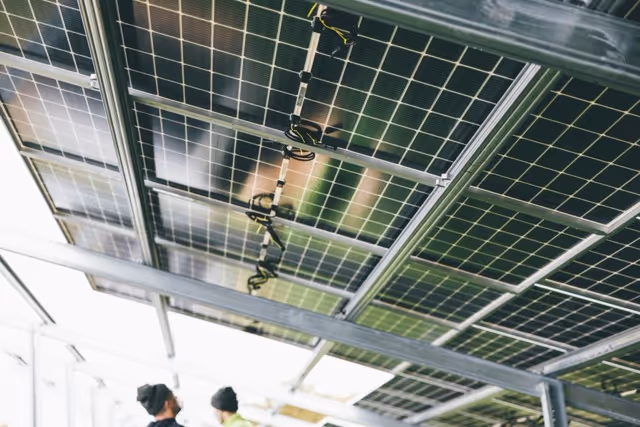 Two people in hats and jackets inspecting solar panels mounted on a metal frame ceiling.