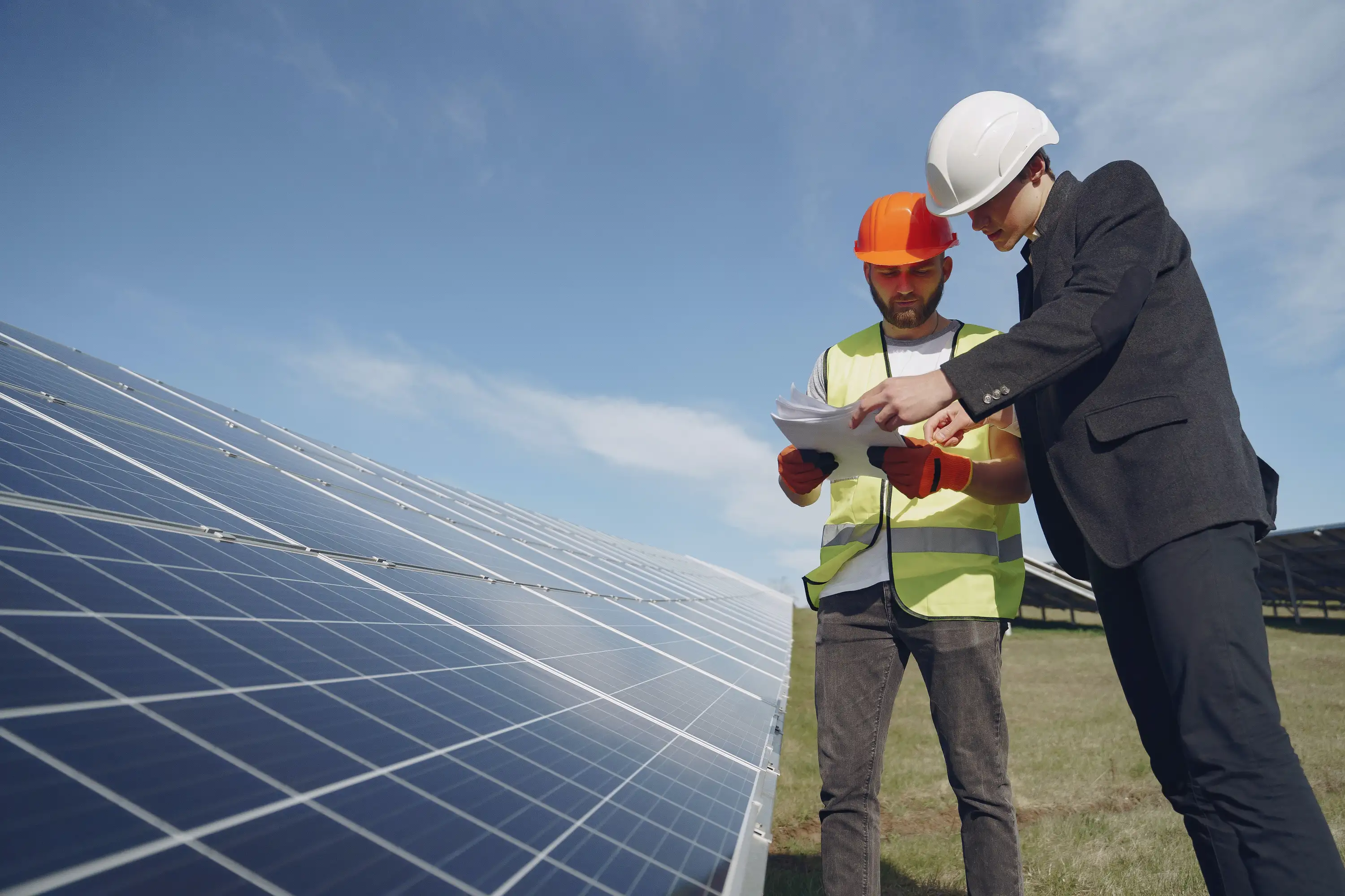 Two workers wearing hard hats and safety gear inspecting documents next to solar panels under a clear sky.